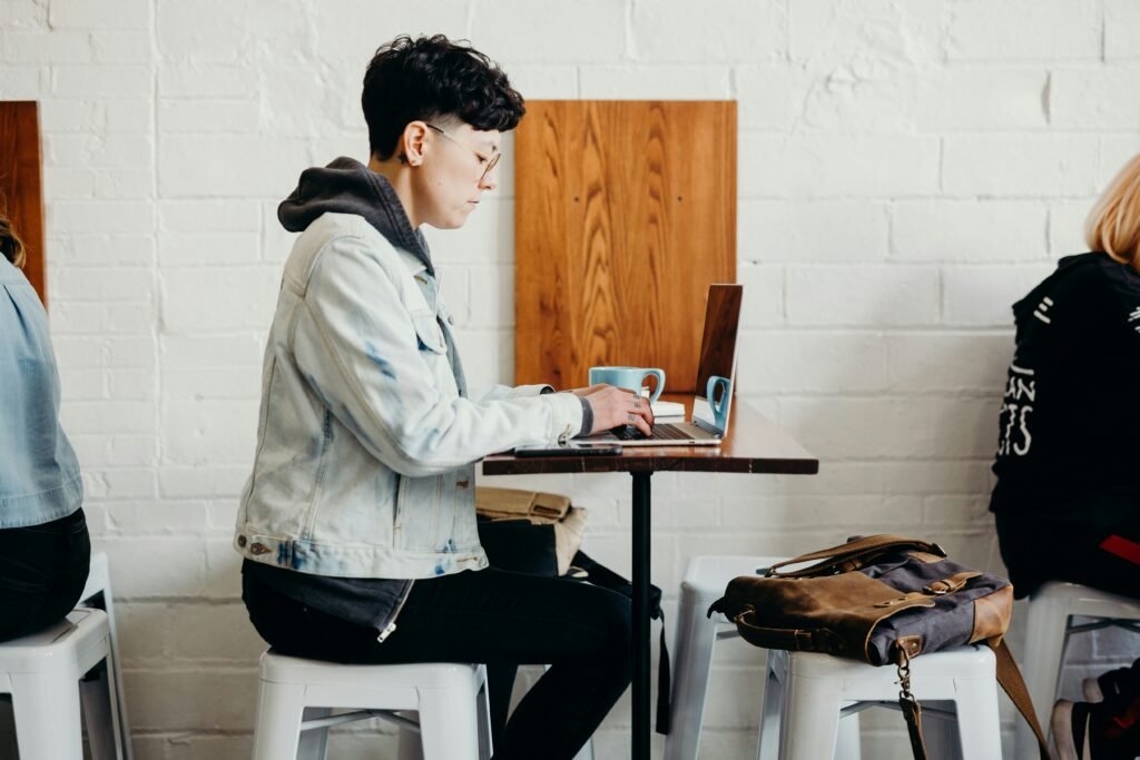 Person with short dark hair wearing glasses and a denim jacket types on a laptop at a cafe table.