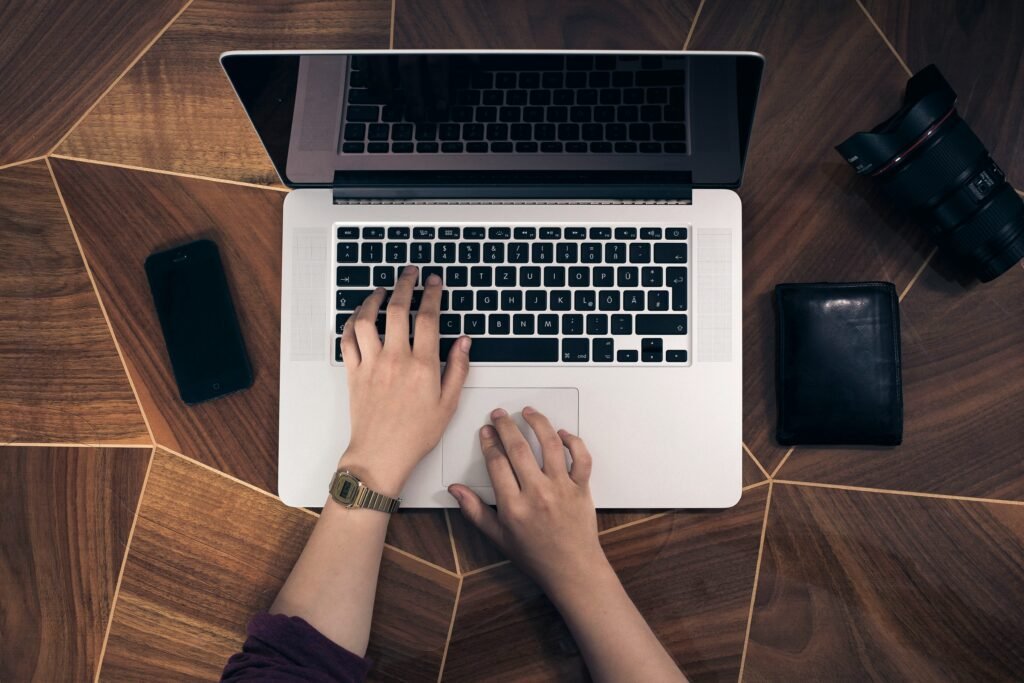 Hands type on a laptop keyboard next to a smartphone, wallet, and camera lens on a wooden desk, showcasing best website tools and products 2025.