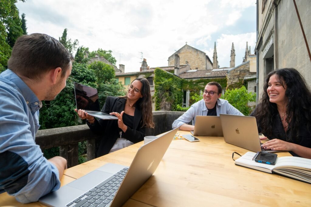 Four people work on laptops at a wooden table on a balcony overlooking buildings.