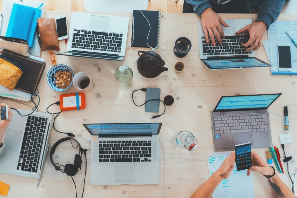 Overhead view of a wooden desk with multiple laptops, phones, and work supplies.