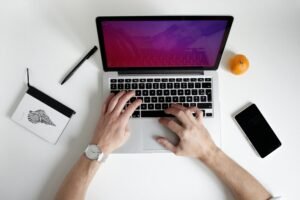 Overhead view of hands typing on a laptop with a notebook, pen, orange, and phone on a white desk.