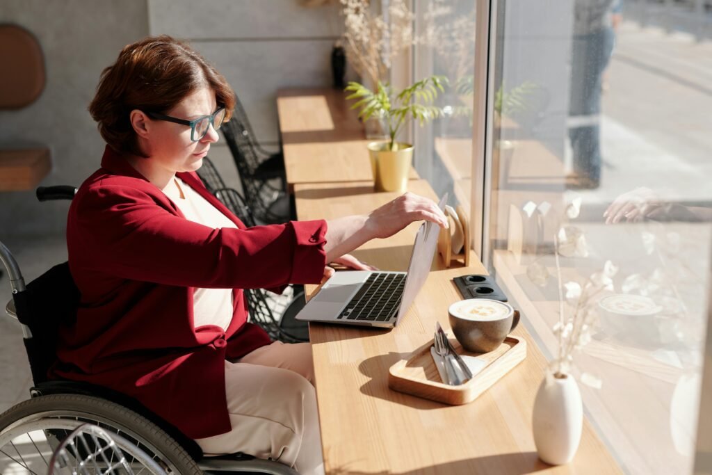 Woman in a wheelchair works on a laptop at a table by a window.