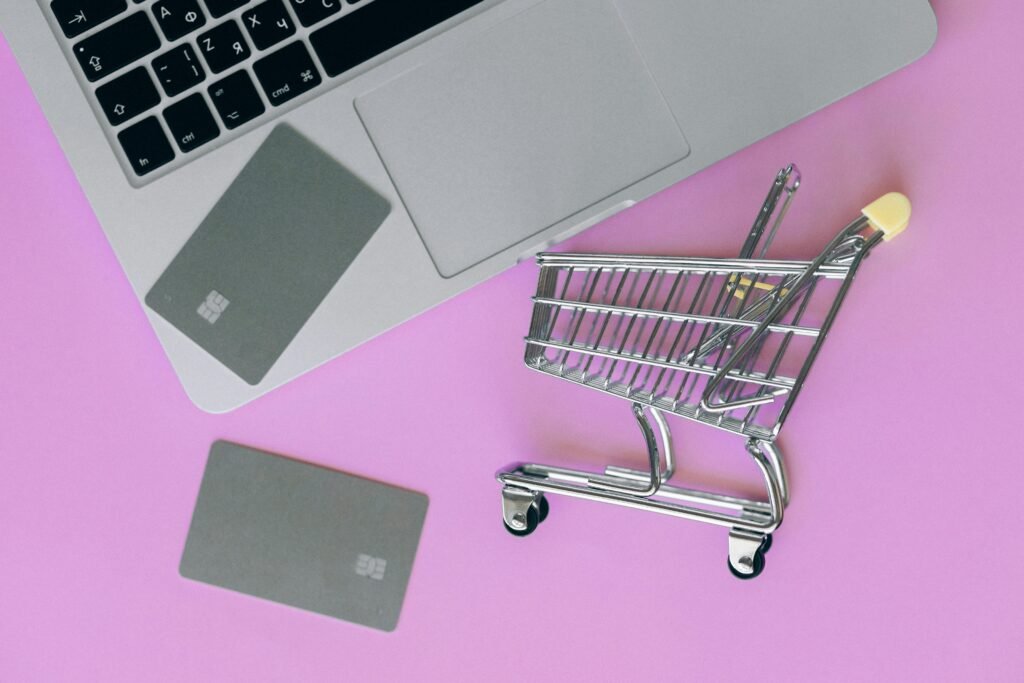 A laptop, credit cards, and a miniature shopping cart are arranged on a pink background.