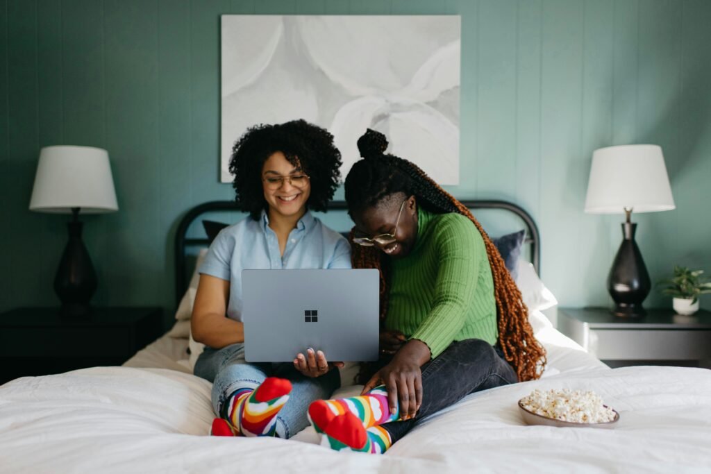 Two women in colorful socks sit on a bed with a laptop and popcorn.