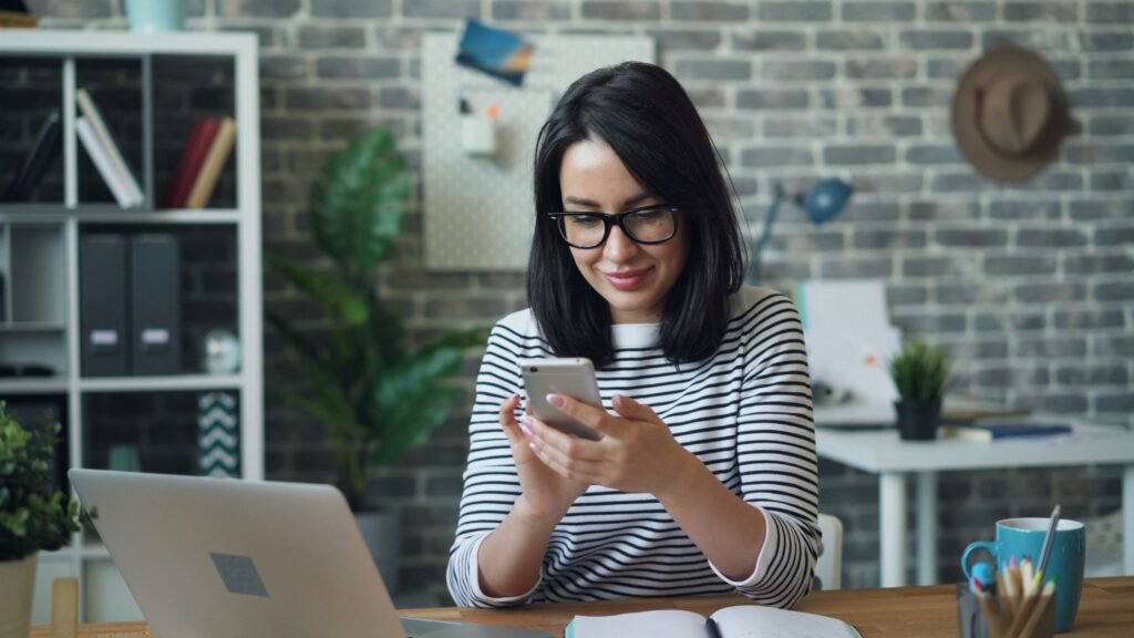 Woman in a striped shirt and glasses smiles while looking at her phone at a desk.