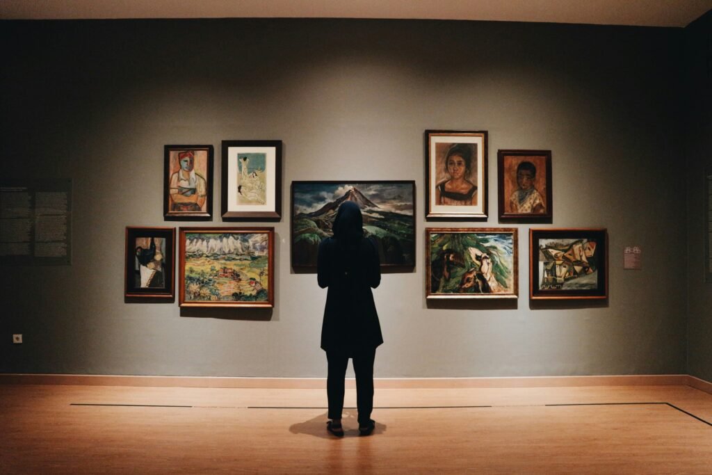 A person in a dark hijab stands before a wall of paintings in a dimly lit art gallery