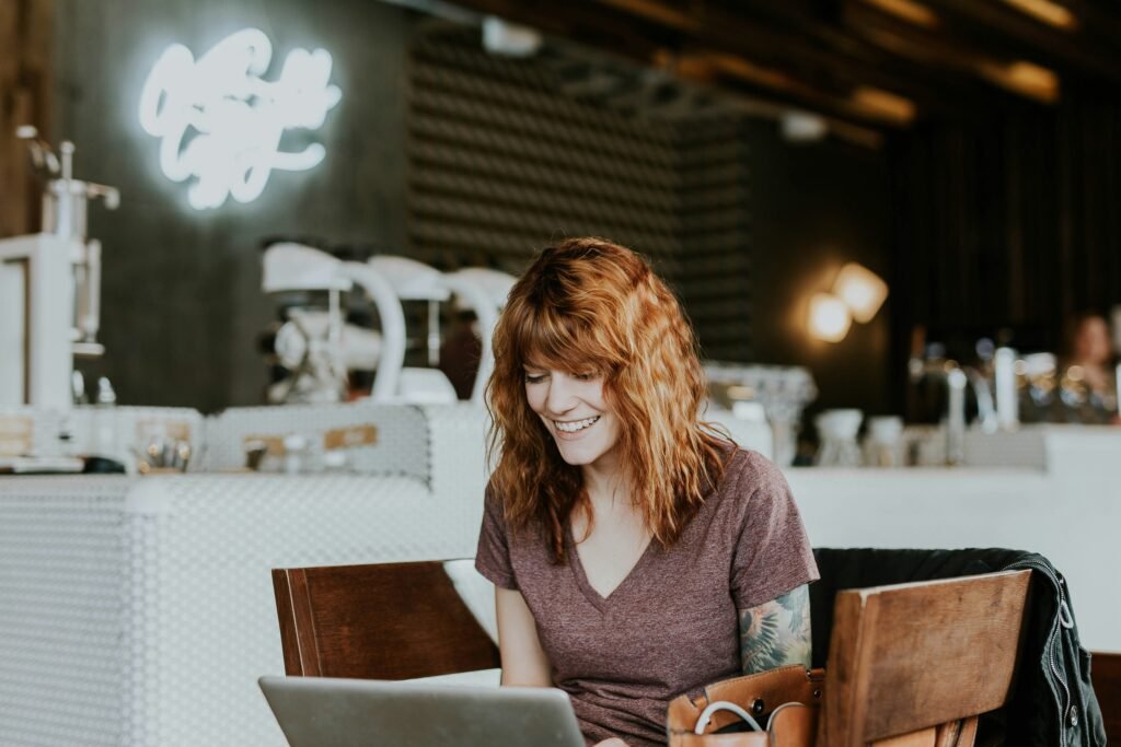 A woman with red hair smiles while looking at her laptop in a cafe