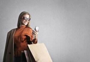 Woman in sunglasses and orange top holds a phone and shopping bags