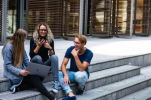 Three young adults sit on outdoor steps, two with a laptop and phone, and one looking at the other two