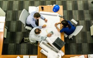 Three people review blueprints and use a calculator at a wooden table