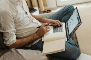 Man in white shirt and jeans types on laptop with notebook open