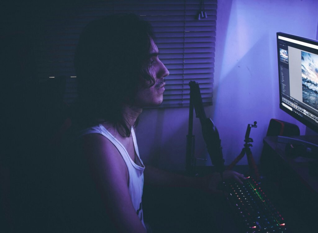 Man in a tank top looks at a computer screen with a microphone and keyboard in a dimly lit room