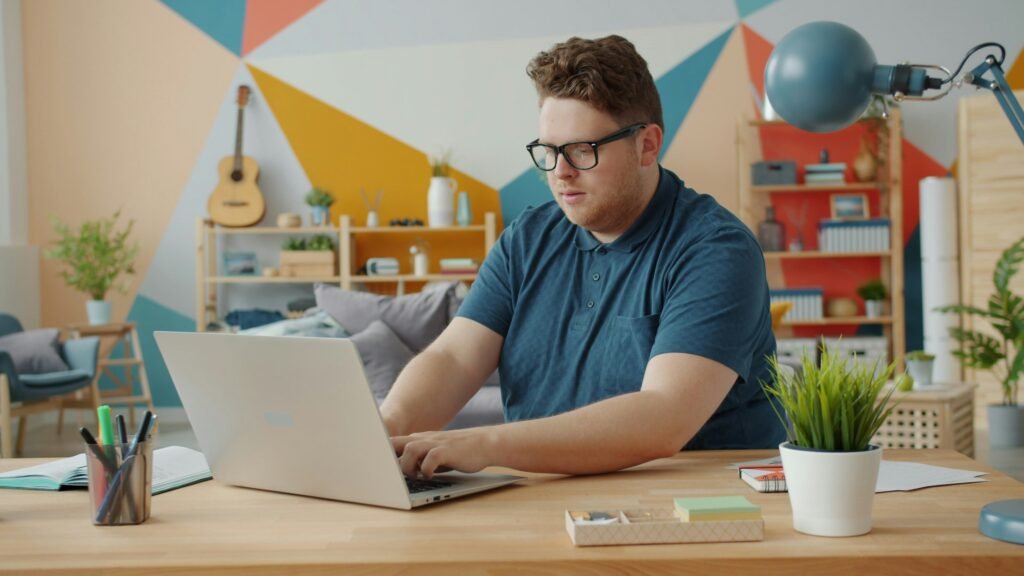 Man with glasses wearing a blue polo shirt works on a laptop at a wooden desk