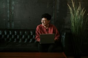 A person with short curly hair and glasses smiles while holding a laptop on a dark leather couch