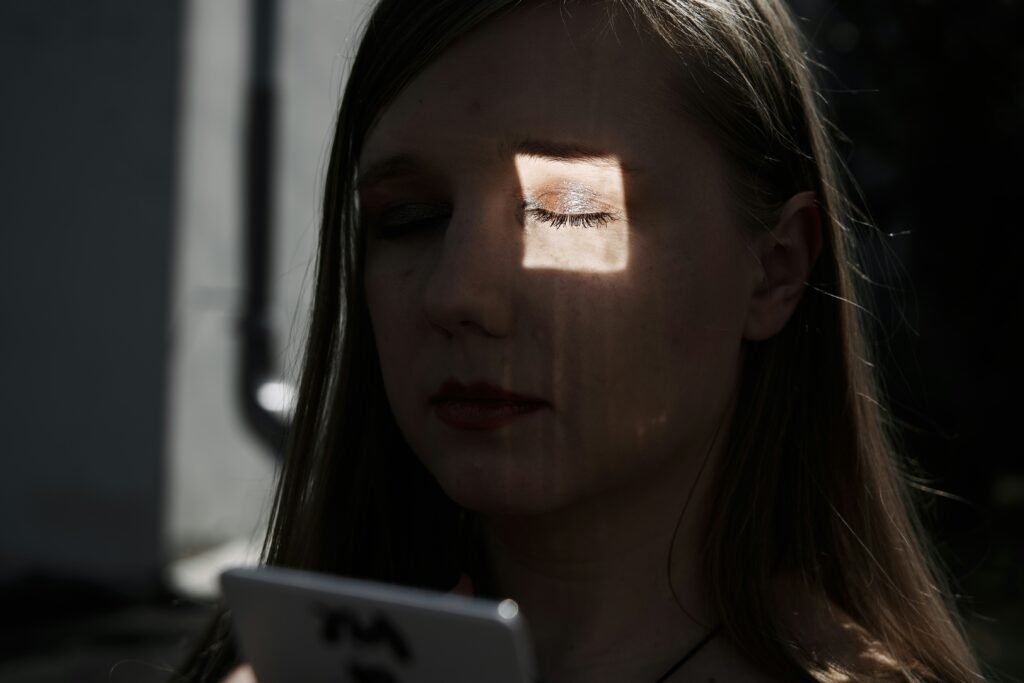 A young woman with closed eyes looks down at a phone, a square of light illuminating her face