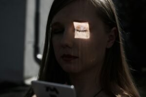 A young woman with closed eyes looks down at a phone, a square of light illuminating her face