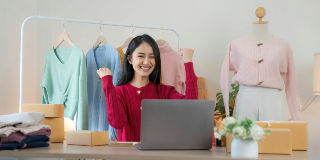 A woman celebrates in front of a laptop with clothing racks and packages