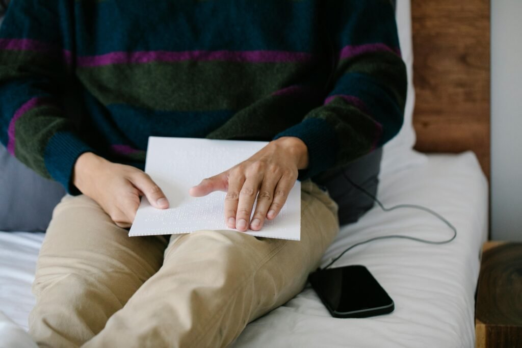 A person reads braille from a white sheet of paper while a smartphone rests on a bed, illustrating ai accessibility