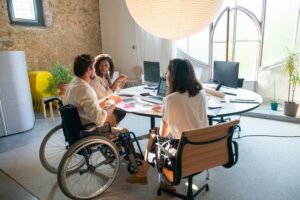 A man in a wheelchair and two women collaborate around a round table with computers and papers