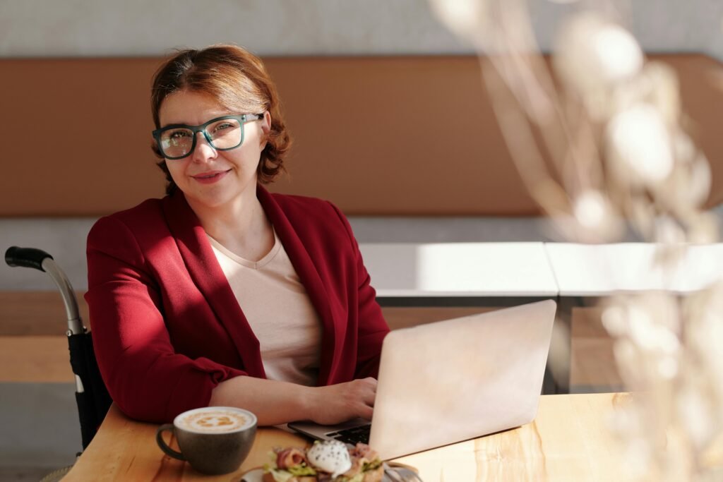 A woman in a wheelchair wearing glasses and a red blazer smiles while working on a laptop at a table with a coffee and a sandwich