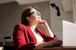 A woman in a red blazer and glasses sits in a wheelchair, looking thoughtfully at a laptop, suggesting a focus on ADA compliant websites