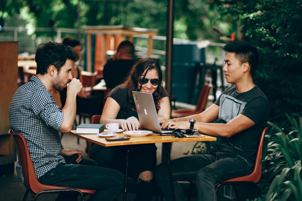 Three people sit at a table outdoors, with a laptop and coffee cups on the table