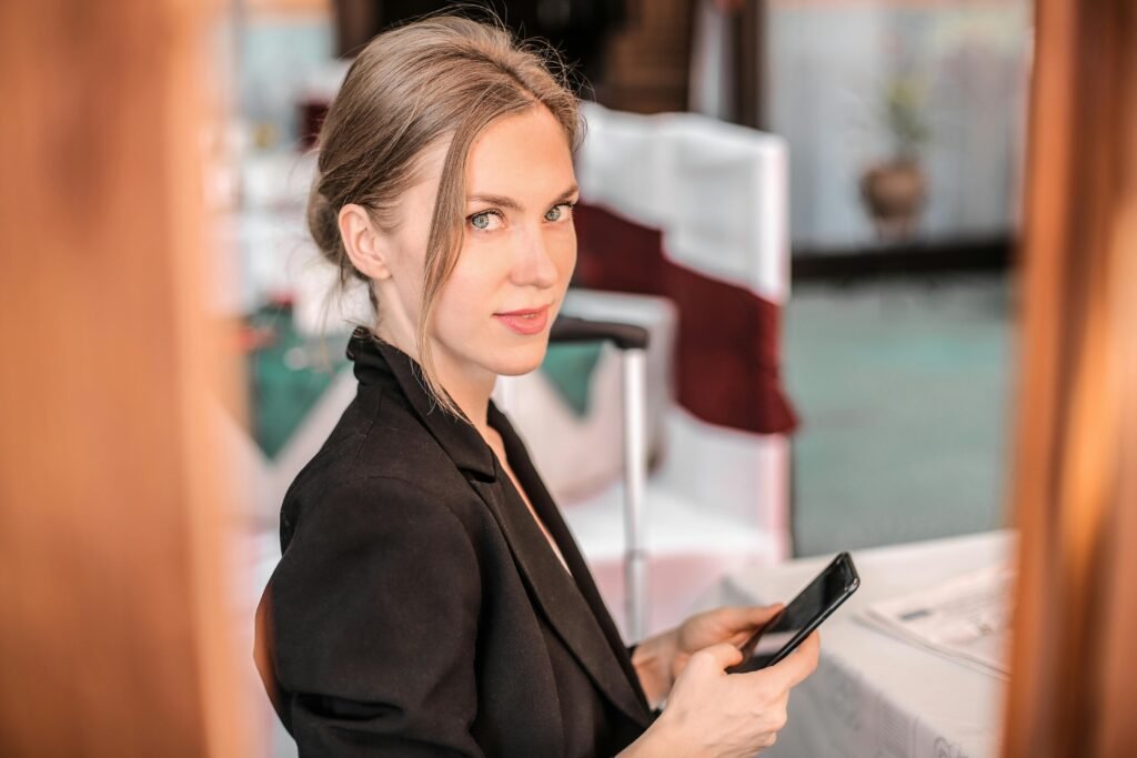 A woman in a black blazer holds a smartphone while looking at the camera, with blurred background elements