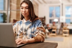 A young woman with dark hair wears a tie-dye hoodie while working on a laptop, illustrating ai model training