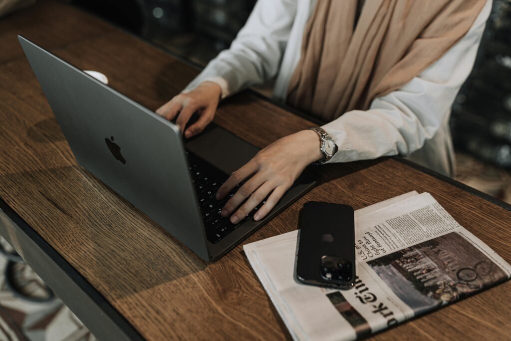 A person wearing a white shirt and tan scarf types on a laptop next to a smartphone and newspaper, possibly representing ai model training