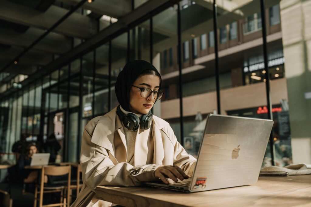A woman wearing a hijab and glasses works on a laptop with headphones around her neck, illustrating ai model training