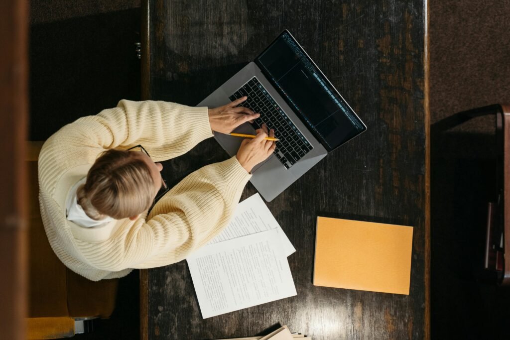 A woman in a cream sweater types on a laptop next to papers and an orange folder, possibly researching image compression tools