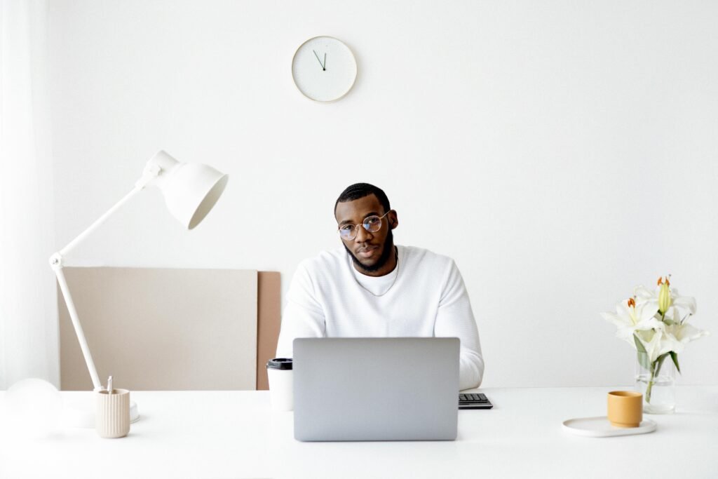 A man in a white sweater and glasses sits at a desk with a laptop, a coffee cup, and a vase of lilies, working with image compression tools