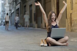 Woman with arms raised in celebration while using a laptop on a city street