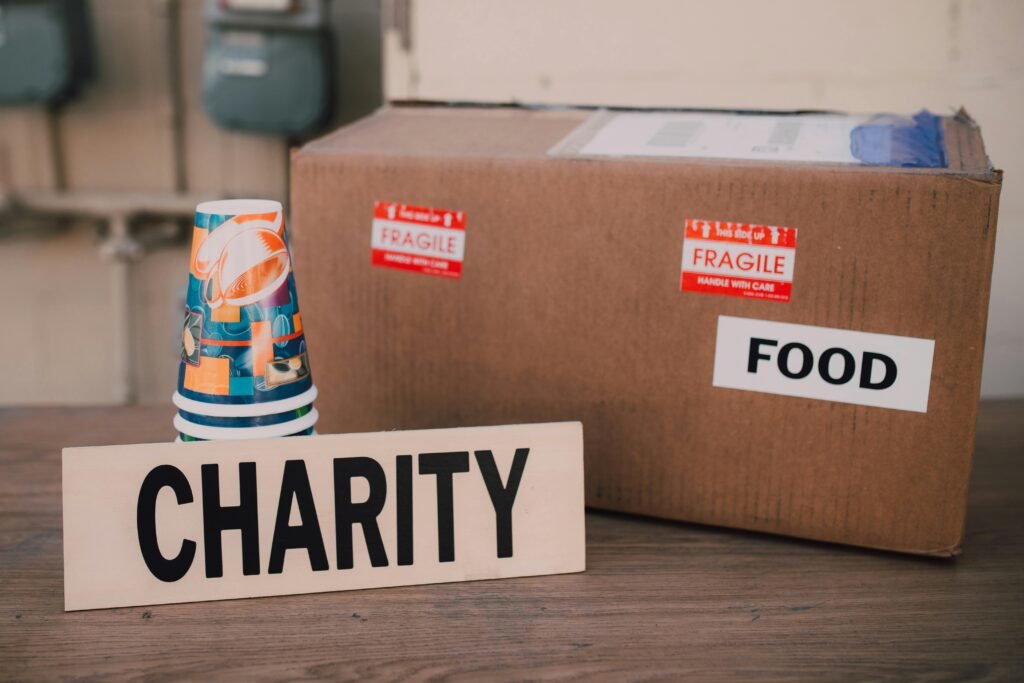 A cardboard box labeled "FOOD" and "FRAGILE" sits next to a stack of colorful cups and a sign that reads "CHARITY"