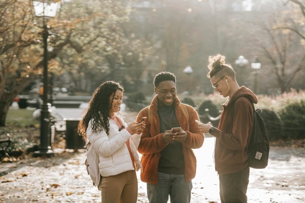 Three young people laugh while looking at their phones outdoors in autumn