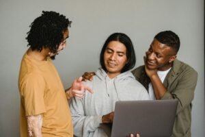 Three men look at a laptop, with one pointing at the screen, as they work online