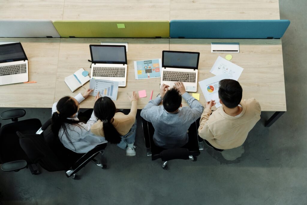 Four people sit at a table with laptops and papers, discussing their work