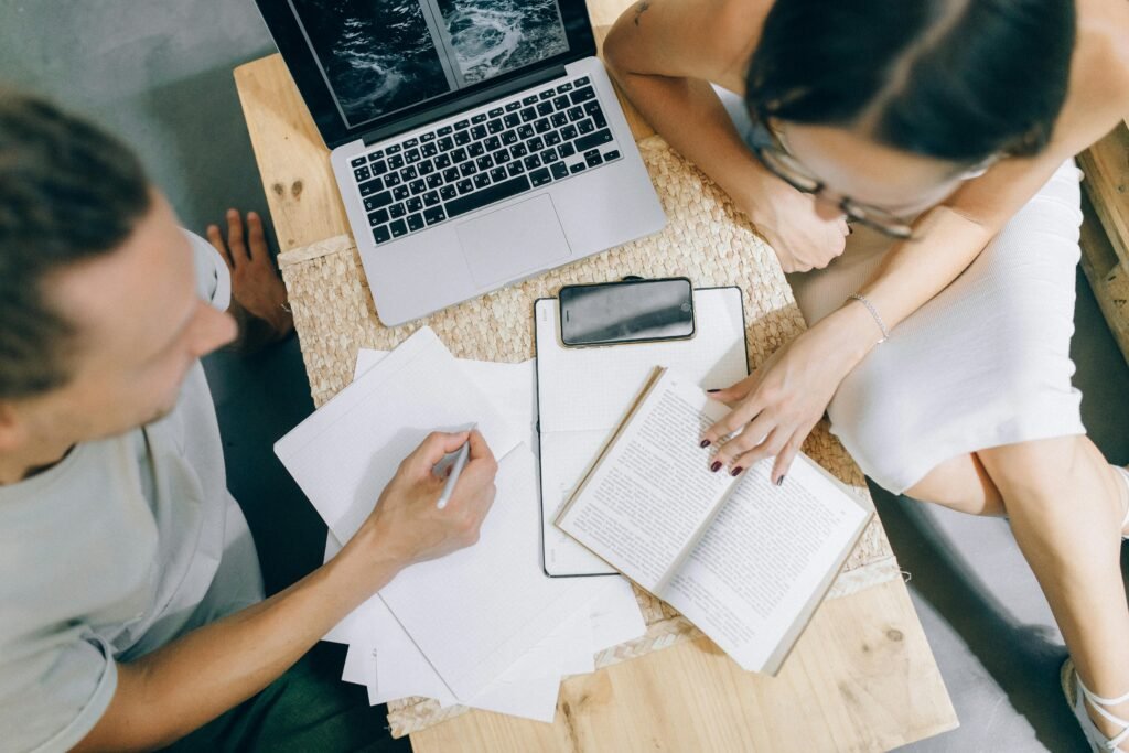 A man and woman study at a wooden table with a laptop, notebook, and book, possibly using a Chrome AI alt text generator
