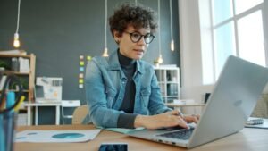 Woman with curly hair and glasses types on a laptop at a wooden desk