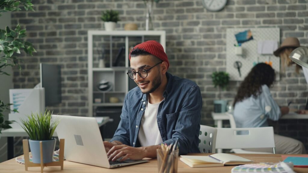 Man in red beanie and glasses works on a laptop at a desk with plants and notebooks