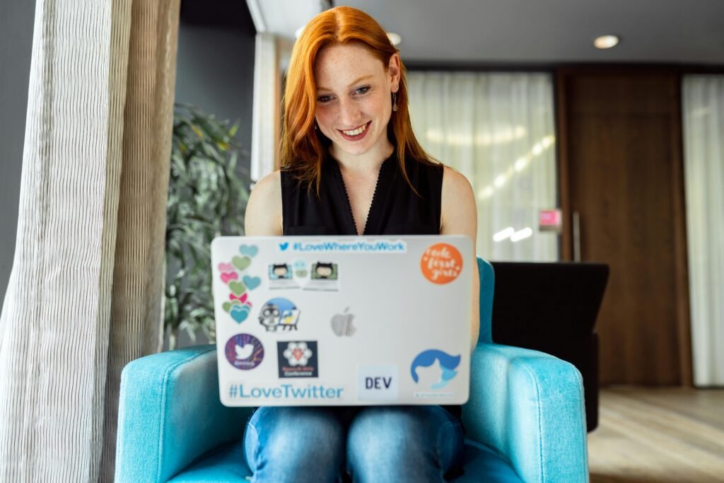 A smiling redhead with freckles works on a laptop covered in stickers