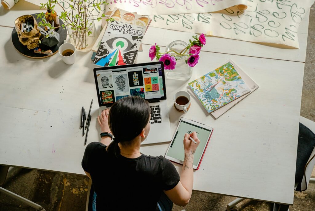 A person works on a laptop and tablet, surrounded by art supplies and magazines, demonstrating the use of AI Alt Text for Publishers