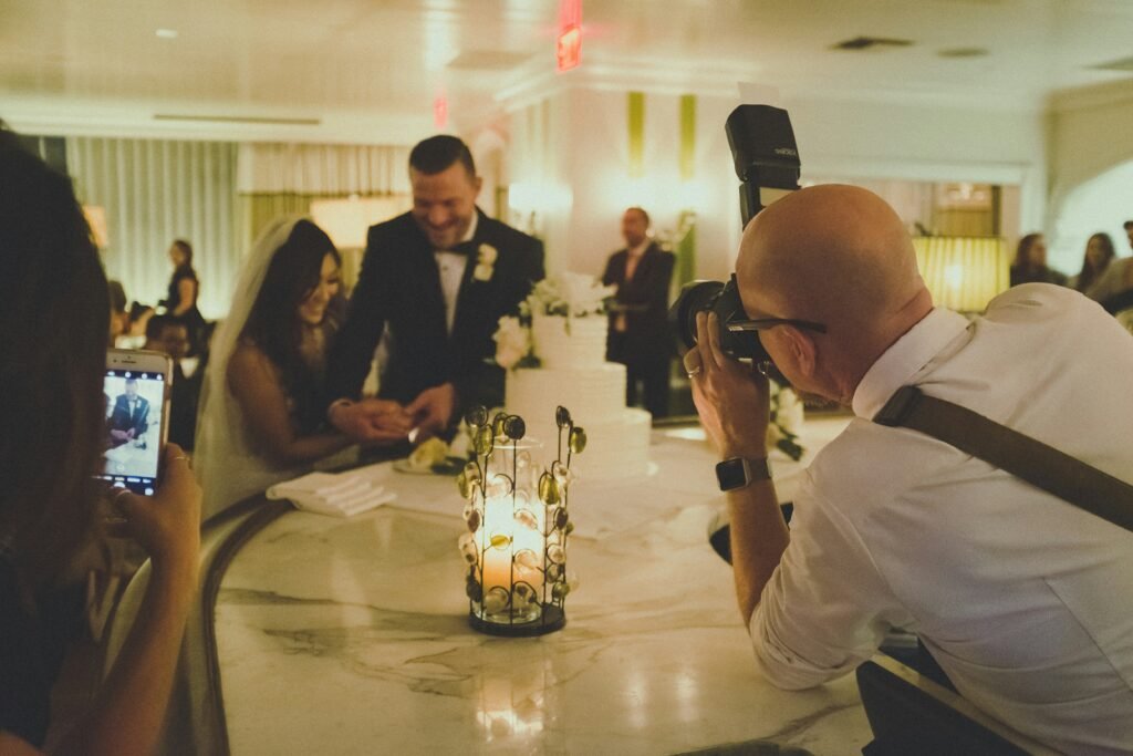 A wedding photographer captures a couple cutting their cake while a guest films the moment on a phone