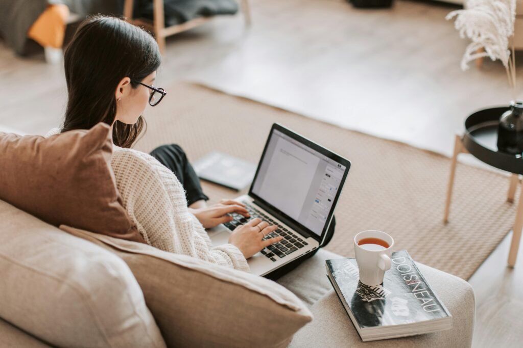 A woman wearing glasses works on a laptop while sitting on a couch with a cup of tea and a book