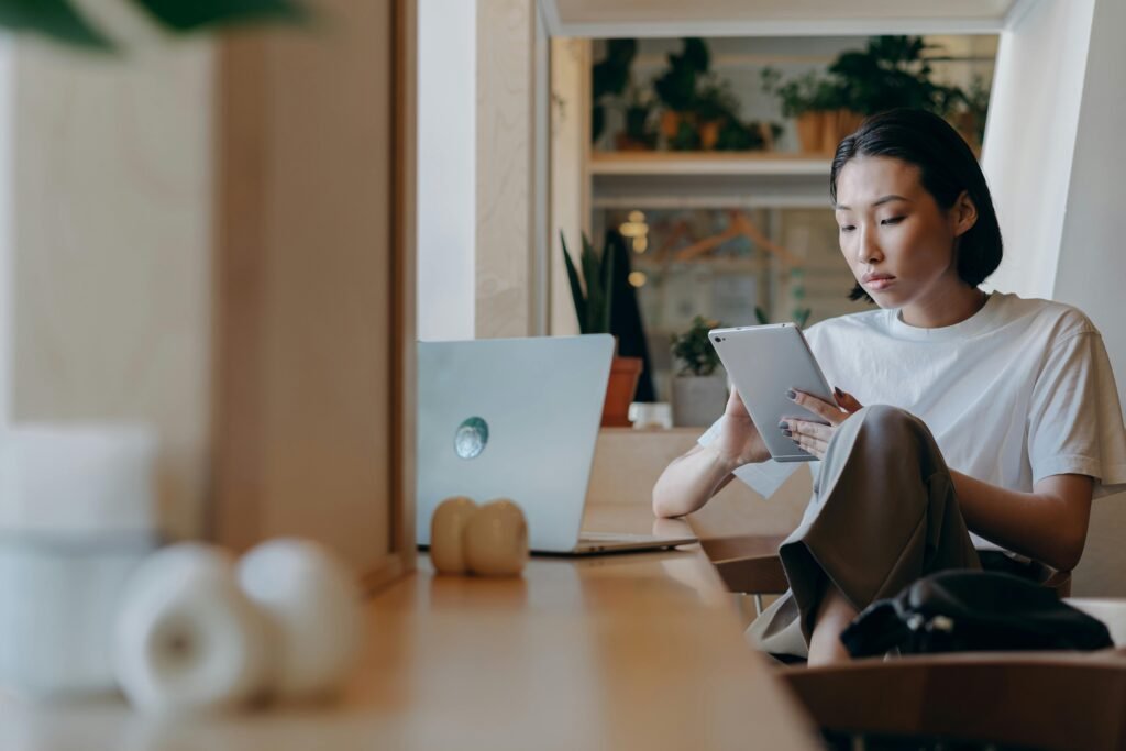 A woman works on a tablet at a wooden desk with a laptop and headphones nearby