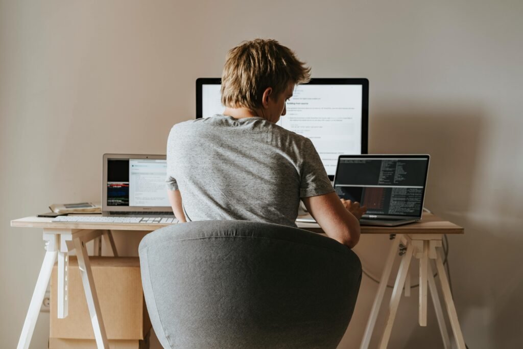A person works on multiple laptops and a desktop computer at a wooden desk