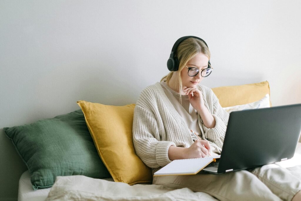 A woman wearing headphones and glasses works on a laptop and notebook while sitting on a bed with pillows