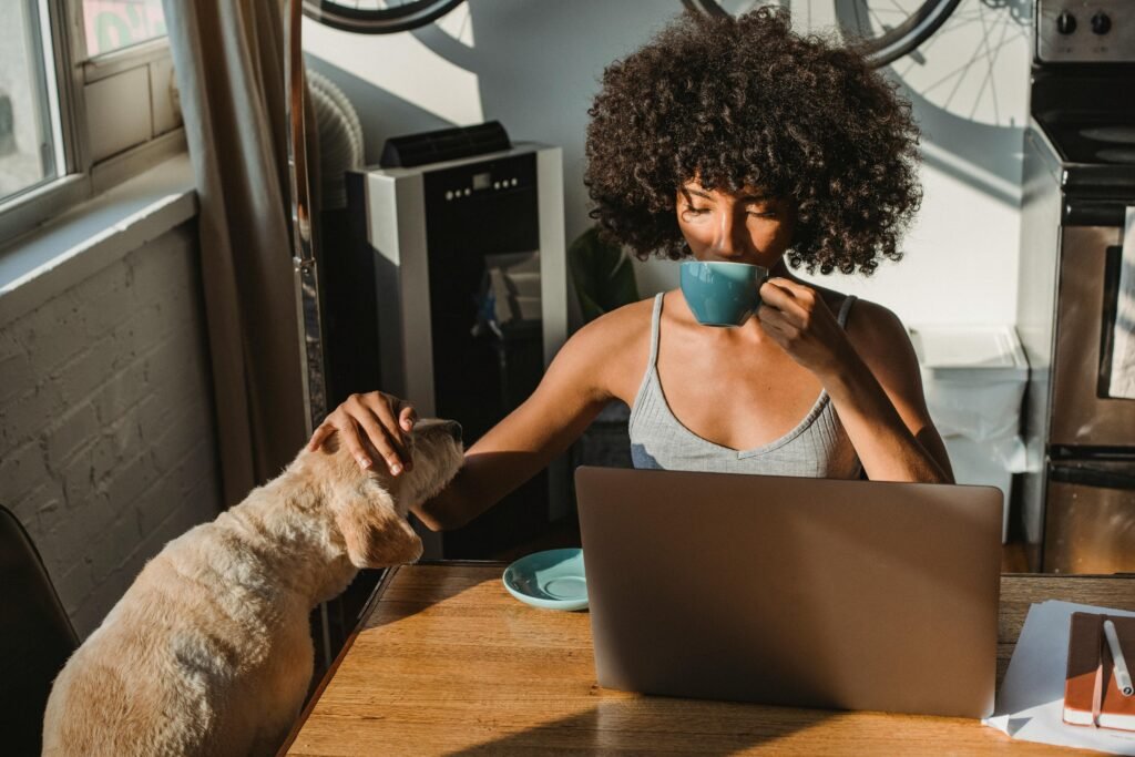 A woman with an afro drinks from a blue mug while petting her dog, with a laptop open on the table
