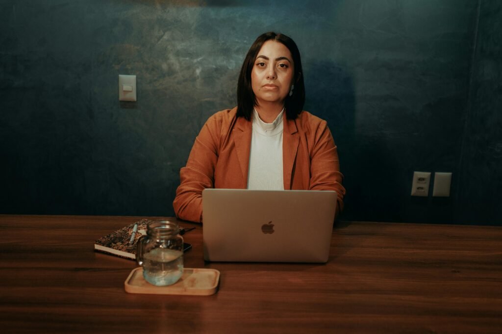 A woman sits at a wooden desk with a laptop, a glass of water, and a notebook, working on wordpress accessibility tools