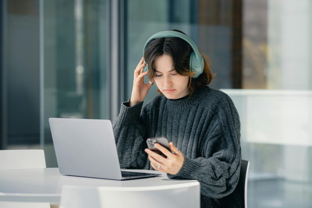 A young woman wearing headphones looks at her phone while sitting at a table with a laptop, possibly using ai alt text generators